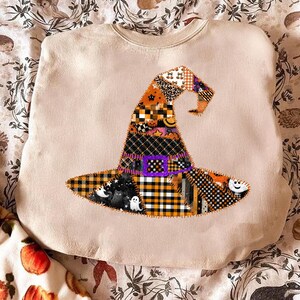 May include: Beige sweatshirt featuring a patchwork witch hat design. The hat is adorned with various patterns, including orange and black plaid, leopard print, and Halloween-themed motifs like ghosts and pumpkins. A purple square buckle is also present.