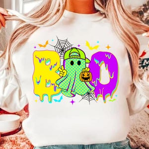 Puede incluir: Sudadera blanca con un gr&aacute;fico colorido de Halloween. El dise&ntilde;o presenta la palabra "BOO" en amarillo y morado, con un personaje fantasma sosteniendo una calabaza. Murci&eacute;lagos y telara&ntilde;as completan el look festivo.