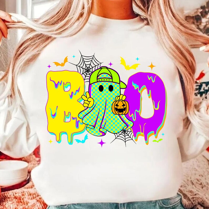 Puede incluir: Sudadera blanca con un gr&aacute;fico colorido de Halloween. El dise&ntilde;o presenta la palabra "BOO" en amarillo y morado, con un personaje fantasma sosteniendo una calabaza. Murci&eacute;lagos y telara&ntilde;as completan el look festivo.