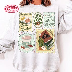 May include: A light gray sweatshirt featuring several illustrated patches with a book theme. Patches include a coffee cup, potted plant, books, and the phrases "Library book card", "Fully Booked", and "One More Chapter".