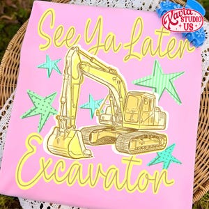 Pu&ograve; includere: T-shirt rosa con un'illustrazione di escavatore giallo e la scritta "See Ya Later Excavator" in un carattere giocoso. Il design include accenti di stelle verdi e blu, creando un look divertente e stravagante. La maglietta ha un contorno giallo chiaro.