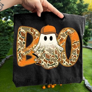 May include: Black t-shirt featuring the word "BOO" in orange with a camouflage pattern. A cartoon ghost wearing a brown baseball cap is in the center. The ghost has black eyes and a camouflage patterned body.