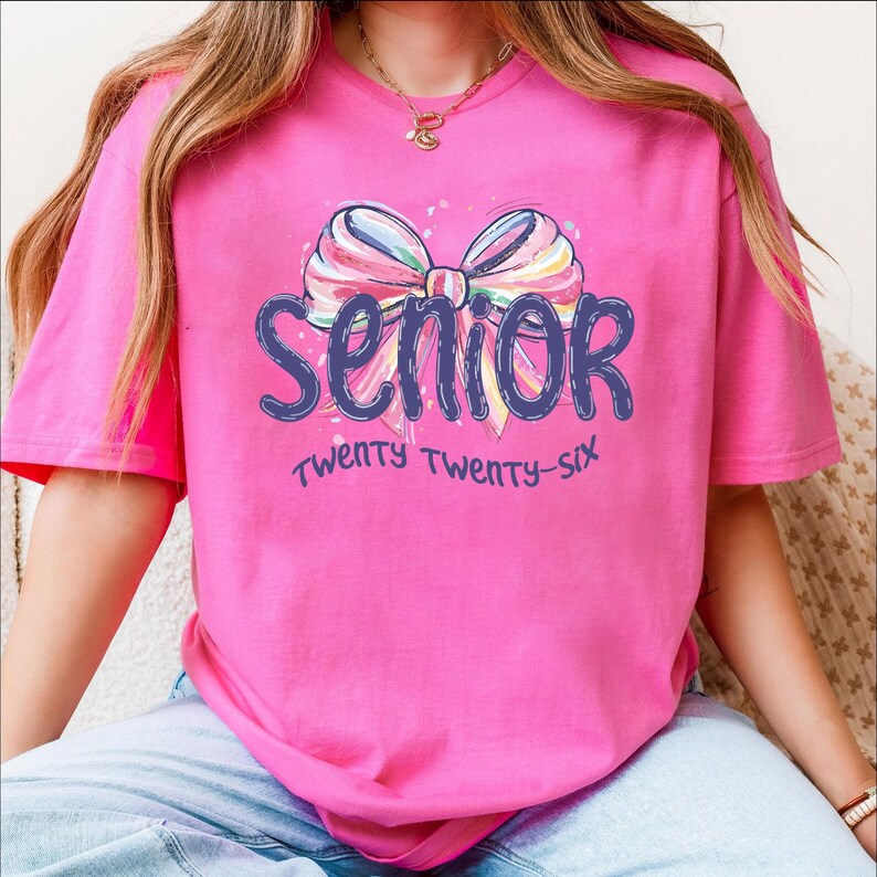 May include: A bright pink t-shirt with the word "Senior" in blue script, adorned with a colorful bow graphic. The text "Twenty Twenty-Six" is below the word "Senior".