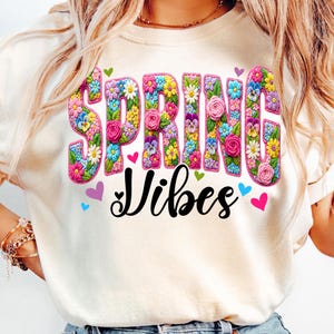 Puede incluir: Camiseta color crema con las palabras "SPRING Vibes" en un dise&ntilde;o floral colorido. La palabra "SPRING" est&aacute; llena de varias flores, y la palabra "Vibes" est&aacute; en una fuente negra. Peque&ntilde;os corazones rodean el texto.