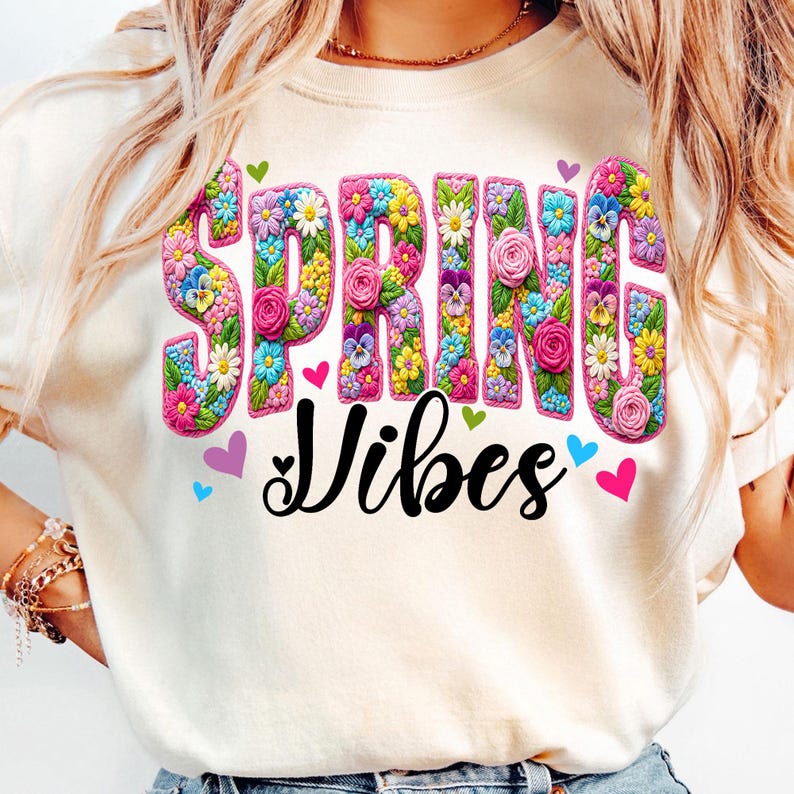 Puede incluir: Camiseta color crema con las palabras "SPRING Vibes" en un dise&ntilde;o floral colorido. La palabra "SPRING" est&aacute; llena de varias flores, y la palabra "Vibes" est&aacute; en una fuente negra. Peque&ntilde;os corazones rodean el texto.