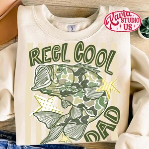 May include: Cream-colored sweatshirt with a green and camouflage fish graphic. The text "REEL COOL DAD" is printed in green. The design includes yellow star accents.
