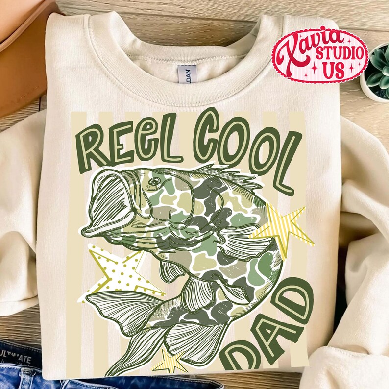 May include: Cream-colored sweatshirt with a green and camouflage fish graphic. The text "REEL COOL DAD" is printed in green. The design includes yellow star accents.
