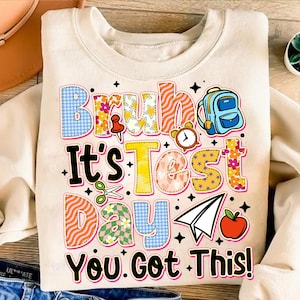 May include: A beige sweatshirt with a colorful graphic that says "Bruh, It's Test Day. You Got This!" The graphic features a backpack, a clock, a paper airplane, an apple, and a sewing needle.