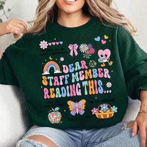 May include: Green sweatshirt with a colorful graphic design that says "Dear Staff Member Reading This..." with a rainbow, butterfly, apple, and other illustrations. The text also says "You are appreciated", "You are respected", and "You are valued".