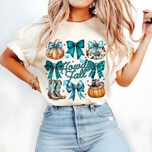 May include: Cream-colored t-shirt featuring a "Howdy Fall" design. The design includes pumpkins, bows, cowboy boots, and a cowboy hat. The bows are teal and leopard print. The pumpkins are orange and decorated with turquoise accents.