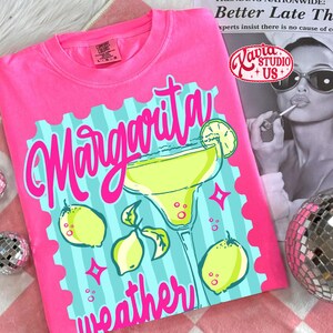May include: A bright pink t-shirt featuring the phrase "Margarita weather" in a fun font. The design includes a margarita glass, lemons, and a light blue background. The shirt also has the "Comfort Colors" brand tag.