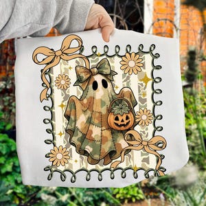 May include: White tote bag featuring a cartoon ghost in a camouflage pattern, holding a pumpkin basket. The design includes bows, flowers, and a decorative border, perfect for Halloween. The overall color scheme is beige, brown, and green.