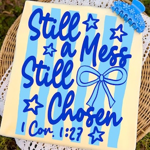 May include: A light yellow t-shirt with blue vertical stripes and the words "Still a Mess Still Chosen" in blue script. The text includes a bow and stars. The Bible verse "1 Cor. 1:27" is also printed on the shirt. A blue hair clip with flower details rests on the shirt.