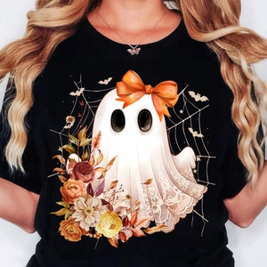 May include: Black t-shirt with a white ghost design. The ghost has an orange bow and is surrounded by a spiderweb and bats. A floral arrangement is at the bottom, creating a Halloween theme.