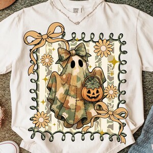 May include: Cream-colored t-shirt featuring a Halloween ghost design. The ghost is draped in a camouflage-patterned sheet and holds a pumpkin basket. The design includes bows, flowers, and a decorative border. The shirt has a Comfort Colors tag.