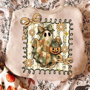 May include: Beige sweatshirt featuring a Halloween ghost design. The ghost is draped in a camouflage pattern and holds a pumpkin basket. The design includes yellow daisies, bows, and a green border, perfect for fall.