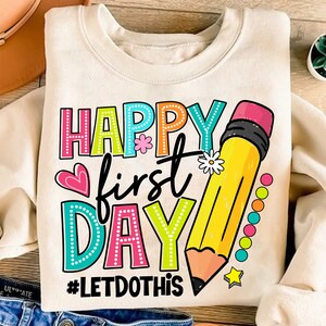 May include: Cream-coloured sweatshirt with the words "HAPPY first DAY" in colourful, dotted letters. A large yellow pencil graphic and the hashtag "#LETDOTHIS" are also featured. Heart and flower accents complete the design.
