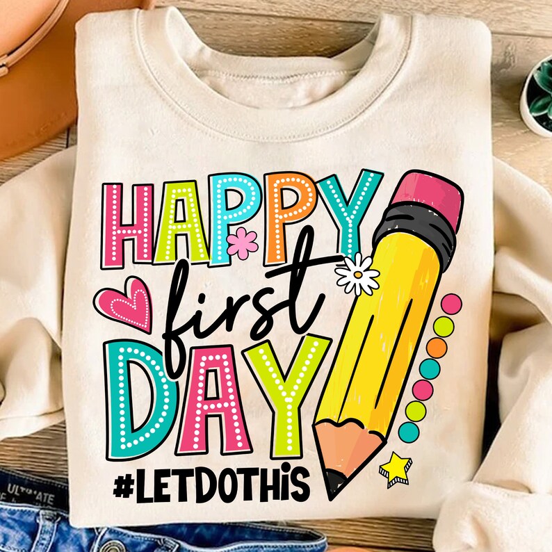 May include: Cream-coloured sweatshirt with the words "HAPPY first DAY" in colourful, dotted letters. A large yellow pencil graphic and the hashtag "#LETDOTHIS" are also featured. Heart and flower accents complete the design.