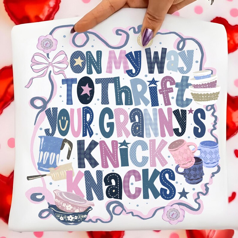 Peut inclure: Taie d'oreiller blanche avec l'inscription "ON MY WAY TO THRIFT YOUR GRANNY'S KNICK KNACKS" dans diff&eacute;rentes polices et couleurs. Le motif comprend des illustrations d'ustensiles de cuisine, d'&eacute;toiles et d'un n&oelig;ud. La taie est tenue par une main avec du vernis &agrave; ongles violet.