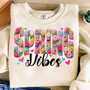 Puede incluir: Sudadera color crema con las palabras "SPRING VIBES" en letras florales grandes y coloridas. Las letras est&aacute;n llenas de varias flores en tonos rosa, amarillo, azul y morado. Peque&ntilde;os corazones de diferentes colores rodean el texto.
