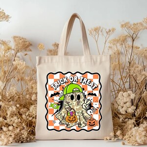 May include: A beige canvas tote bag featuring a Halloween-themed graphic. The graphic includes a cartoon ghost wearing a green cap, holding a pumpkin, and giving a peace sign. The words "TRICK OR TREAT" are displayed above the ghost on a checkered orange and white background.