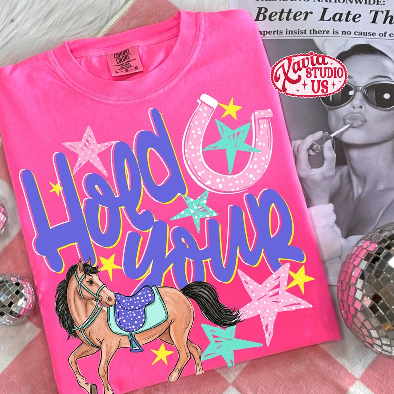 May include: A bright pink t-shirt with the words "Hold Your" in blue script, a horseshoe, stars, and a horse graphic. The shirt also features the text "Kavia Studio + US+".