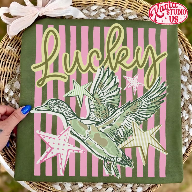 May include: Olive green t-shirt with a duck graphic and the word "Lucky" in a whimsical font. The design includes pink and green striped background and star accents. The shirt is a casual, everyday wear item.