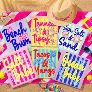 Puede incluir: Toallas de playa con textos coloridos y gr&aacute;ficos tem&aacute;ticos de playa. Las toallas presentan frases como "Beach Bum", "Tanned & Tipsy" y "Sun, Salt & Sand". Tambi&eacute;n se ven un sombrero de paja y gafas de sol, lo que sugiere un tema de playa o vacaciones.