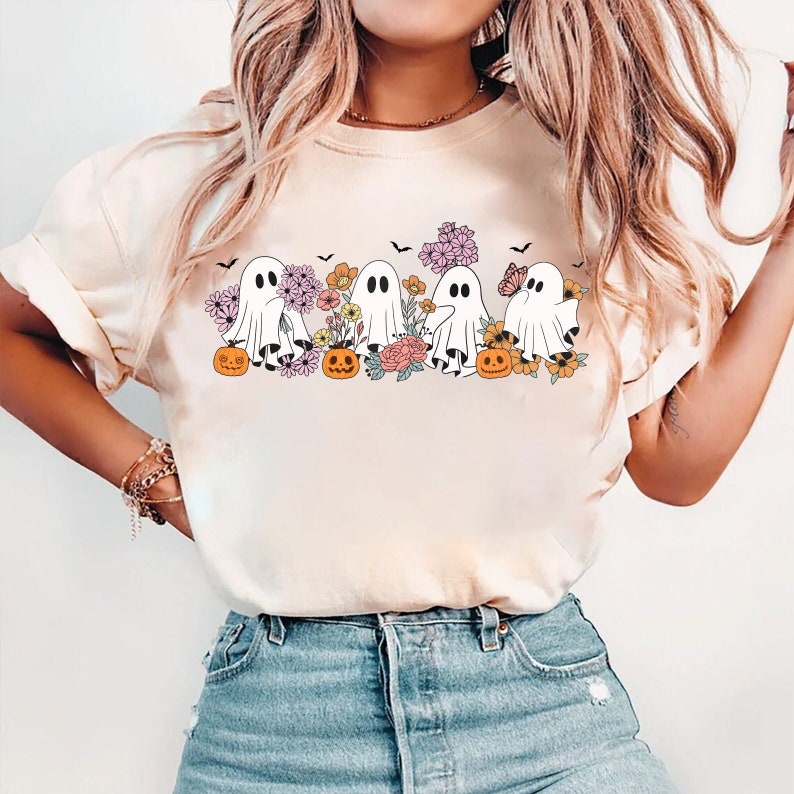 May include: A white t-shirt with a graphic of five ghosts in a line, each holding a jack-o-lantern. The ghosts are surrounded by flowers and butterflies. The shirt is a light cream color.