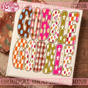 May include: A white t-shirt featuring the text GOBBLE GOBBLE in large, colorful, patterned letters. The letters display various designs including polka dots, plaid, checkered patterns, and stars in shades of brown, orange, pink, and green.