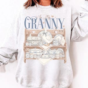 May include: A light gray sweatshirt featuring a graphic of a wooden shelf displaying vintage dishware, a goose, and the text "In My Granny Era" in blue script. The design includes hearts and a chicken figurine.