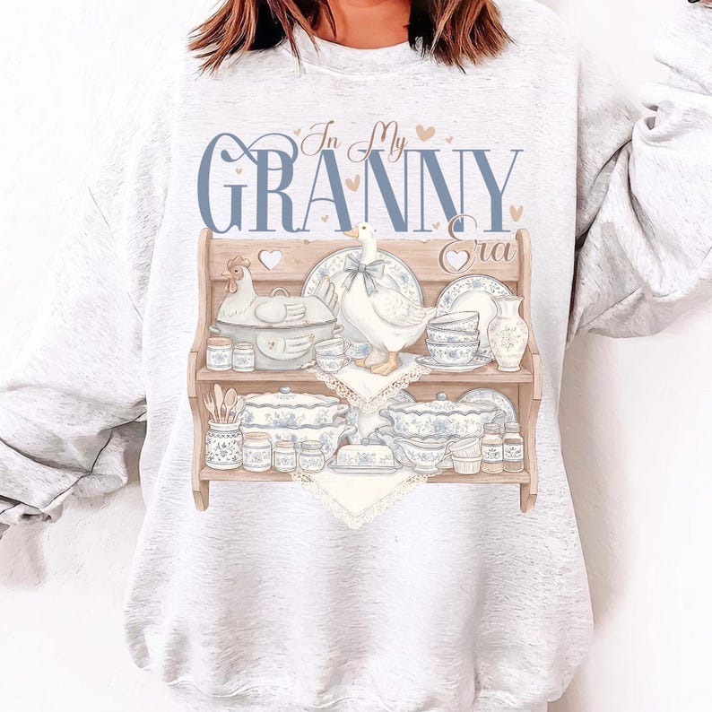 May include: A light gray sweatshirt featuring a graphic of a wooden shelf displaying vintage dishware, a goose, and the text "In My Granny Era" in blue script. The design includes hearts and a chicken figurine.