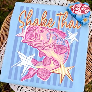 May include: Light blue t-shirt with a pink fish illustration and the text "Shake That" in red and yellow. The design includes white and yellow stars. The shirt has vertical blue stripes.