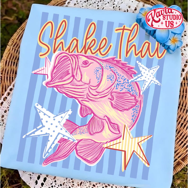 May include: Light blue t-shirt with a pink fish illustration and the text "Shake That" in red and yellow. The design includes white and yellow stars. The shirt has vertical blue stripes.