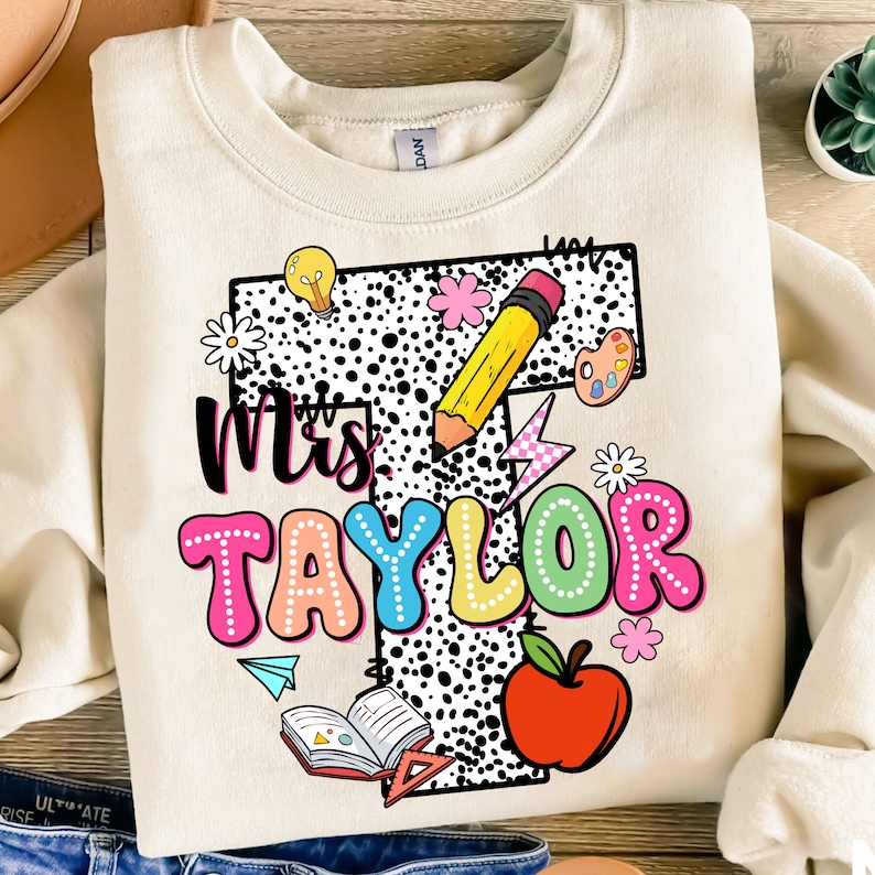 May include: A beige sweatshirt with a black and white polka dot background. The text "Mrs. Taylor" is in colorful bubble letters. The letter "T" is decorated with a pencil, a lightbulb, a flower, a paint palette, a lightning bolt, and a book. There is also a red apple and a paper airplane.