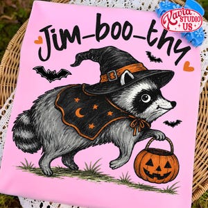 May include: A light pink t-shirt featuring a graphic of a raccoon wearing a black witch hat and a black cape with orange stars and a crescent moon. The raccoon holds an orange jack-o'-lantern bucket. The text reads Jim-boo-thy in black script.