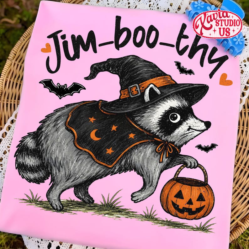 May include: A light pink t-shirt featuring a graphic of a raccoon wearing a black witch hat and a black cape with orange stars and a crescent moon. The raccoon holds an orange jack-o'-lantern bucket. The text reads Jim-boo-thy in black script.