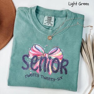 May include: Light green t-shirt with the word "Senior" in purple script and a colorful bow graphic. The text "Twenty Twenty-six" is below the word "Senior". A gold necklace with a pendant is on the shirt.