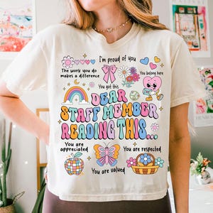 May include: A white t-shirt with a colorful graphic design that says "Dear Staff Member Reading This..." with illustrations of a rainbow, butterfly, peace sign, and flowers. The text also says "You are appreciated, You are respected, You are valued".