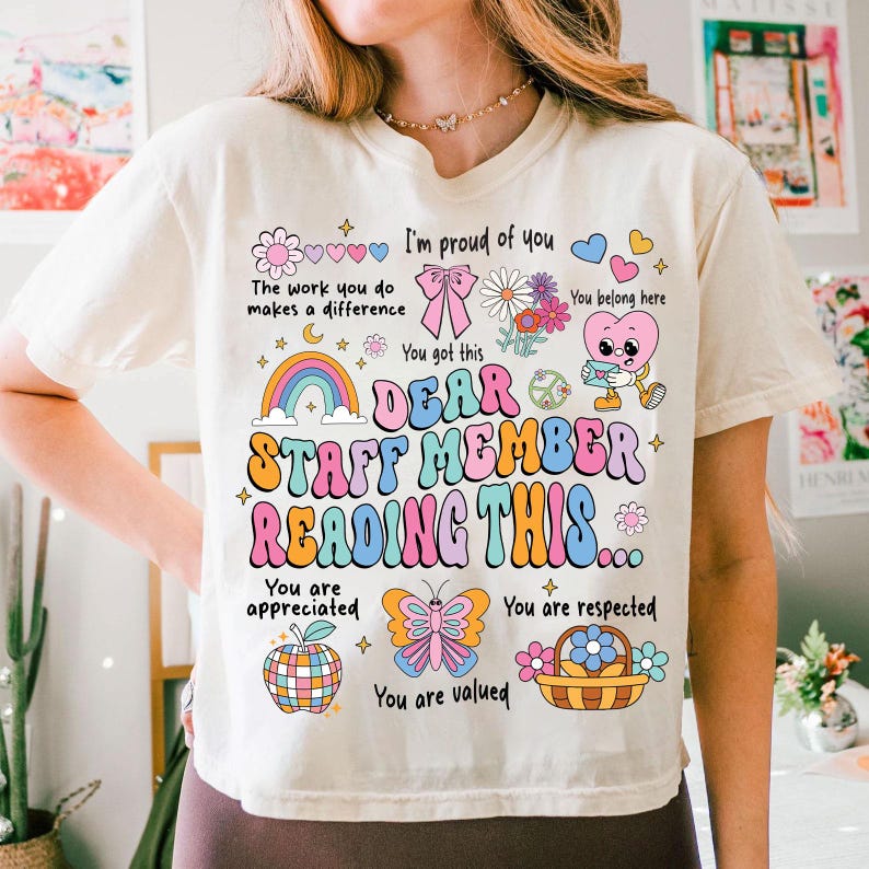 May include: A white t-shirt with a colorful graphic design that says "Dear Staff Member Reading This..." with illustrations of a rainbow, butterfly, peace sign, and flowers. The text also says "You are appreciated, You are respected, You are valued".