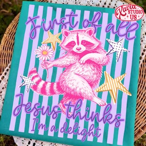 May include: A teal and lavender striped decorative towel featuring a pink raccoon holding a daisy. The text reads "First of all Jesus thinks I'm a delight" in purple script. Gold and white stars are scattered around the raccoon.
