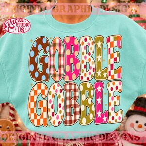 May include: A light blue crewneck sweatshirt featuring the text GOBBLE GOBBLE in large, multi-patterned letters. The letters display various designs including polka dots, plaid, checkered patterns, and stars in orange, pink, brown, and green.
