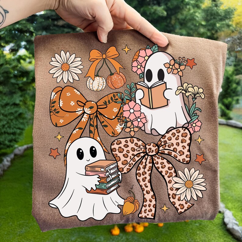 May include: Brown t-shirt featuring a whimsical design with two cartoon ghosts, bows, flowers, pumpkins, and stars. One ghost reads a book, the other holds a stack of books. The bows have floral and leopard print patterns.