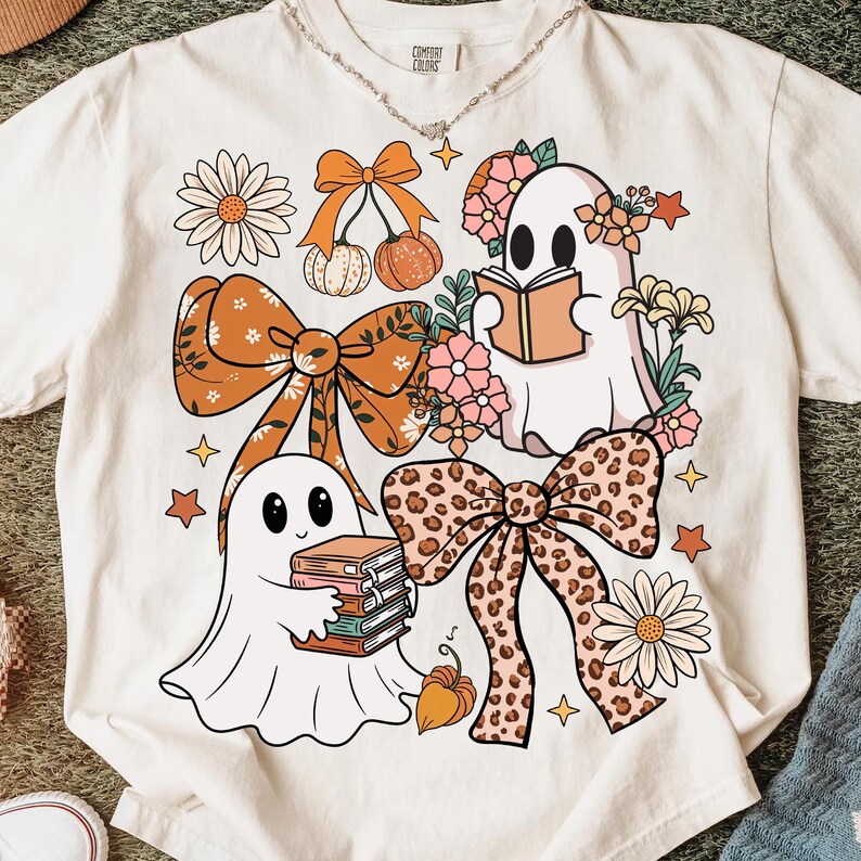 May include: Cream-colored t-shirt featuring a whimsical design with two cartoon ghosts, bows, flowers, and pumpkins. One ghost reads a book, while the other holds a stack of books. The bows have a leopard print and orange color.