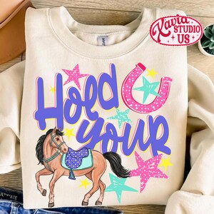 May include: Cream-colored sweatshirt with the text "Hold Your" in purple script, a pink horseshoe, and stars. A brown horse with a blue saddle is featured below the text. The design includes pink, yellow, and turquoise accents.