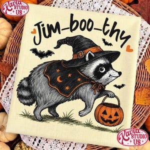 May include: A cream-colored fabric featuring a black and white illustration of a raccoon wearing a black witch hat with an orange band and a black cape with orange stars and a crescent moon. The text Jim-boo-thy is printed above with orange hearts.