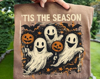 May include: Brown fabric with a Halloween graphic. The design shows three smiling ghosts, jack-o'-lanterns, bats, and stars. The text "'TIS THE SEASON" is printed at the top. The fabric is held up by a hand.