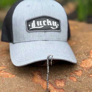 May include: A gray baseball cap with a black mesh back. The cap features a rectangular patch with the word "Lucky" in white script. A silver metal hat clip is attached to the brim.