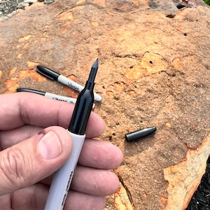 Puede incluir: Un marcador permanente negro con una etiqueta blanca que dice "Permanent Marker". El marcador se sostiene en una mano contra un fondo marrón texturizado.