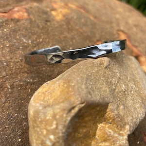 May include: A silver hammered metal bracelet with a smooth, polished finish. The bracelet is open and has a slightly curved shape.